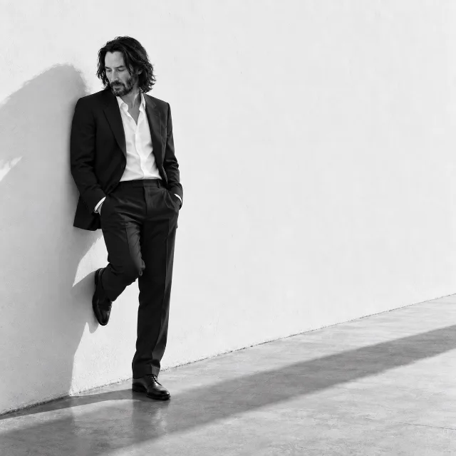 Unofficial AI-generated image of Keanu Reeves: black suit and white shirt, leaning in a bright empty space with a long shadow.
